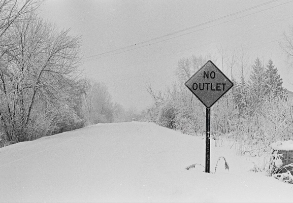 A black-and-white photo of a snow-covered road disappearing into the distance. A roadside sign reading “NO OUTLET” stands on the right, surrounded by trees heavily coated in snow. The scene feels quiet, cold, and slightly absurd in its simplicity