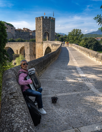 Color photo of a seated man playing the accordion at the entry to a medieval stone bridge with the bridge and its stone tower in the background. 