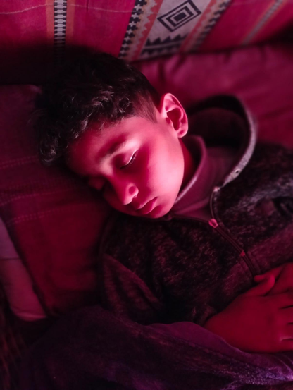 Saji is asleep, resting his head on a pillow under red lighting (because of the red fabric of the tent), wearing a hooded zip-up jacket.
