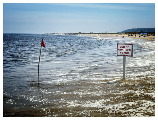 A beach under a blue sky with streaks of high clouds. In the foreground are a small red warning flag on a spindly pole and a signpost that reads “END OF GUARDED BEACH.” Both flagpole and post are partly submerged by a surge of incoming waves. In the distance is the dry shore, dotted with tiny people and beach umbrellas. At the horizon is a line of trees and a low hill in silhouette.