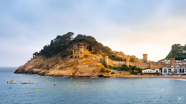Color photo of a rocky coastal hill extending into the Mediterranean Sea that is covered with the walls and towers and buildings of an old medieval town. 