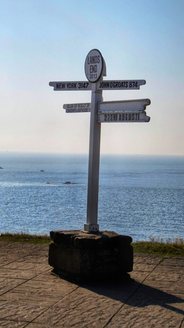 This image shows a wooden signpost situated by the sea, set against a scenic coastal backdrop. The signpost is labelled "Lands End 2013" at the top. It features several directional signs pointing towards various destinations: "New York 3147," "John O'Groats 874," "Isles of Scilly 28," and "Lizard Lighthouse 11." There is also a sign indicating the date "27th August." The signpost is mounted on a stone base and stands on a paved area, with the ocean and a clear sky visible in the background. The overall scene evokes a sense of adventure and the beginning or end of a journey.