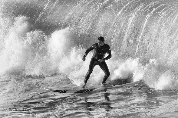 This is a black and white water sports photo in landscape format of a man surfing ahead of a large and very angry breaking wave. Hermosa Beach, California (2016).

In the centre of the image is a young to middle-aged male surfer, wearing a black wetsuit and standing on his white and black surfboard. He is facing the camera while his board is heading diagonally across the image from right to left and heading towards the lower left corner. The water he is crossing is the fairly calm area ahead of the breaking wave behind him. Immediately behind him is a small zone of brilliant white, steaming surf that becomes more dense and taller further to the right, where it touches the upper right corner of the image. Behind the surf is the tall wall of angry water of the breaking wave that dominates the background of the photo. The part of the wave in view is about twice the height of the surfer, so say about 4 metres. The breaking wave looks more like the water cascading over a large water fall or weir, rather than a wave as the pacific ocean falls at a near vertical angle behind our surfer.