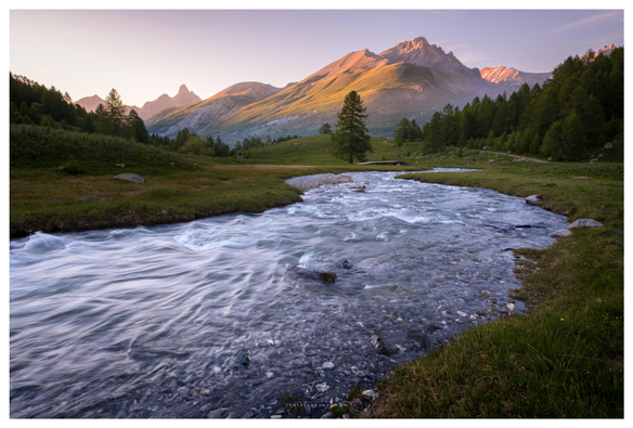 A river flows through a flat and green valley. On the horizon stretches a mountain whose meadows and stones are bathed in an orange light by the setting sun.