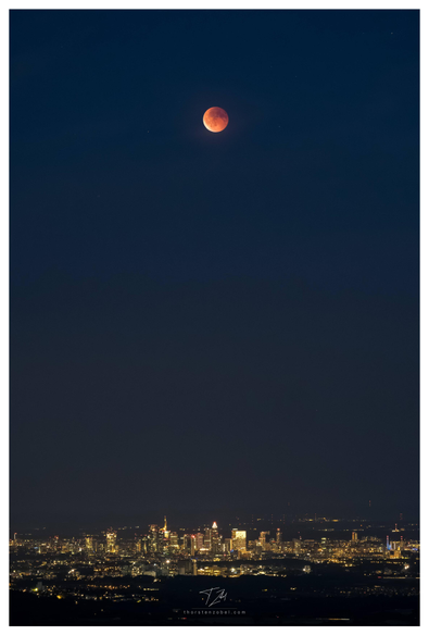 A vertically taken photo showing the blood moon high above over Frankfurt's illuminated skyline.