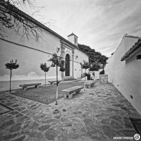 A black and white photo of a quiet, narrow plaza lined with small trees and stone benches. On the left is the Santo Domingo de Guzmán church with a bell tower. Soft, overcast sky adds to the serene atmosphere.