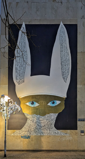 A striking mural painted on an outdoor wall, depicting a stylised, abstract face with rabbit-like ears. The ears are elongated, white, and adorned with intricate black text. The face itself features large, vivid blue eyes and a golden, mask-like covering over the nose and cheeks. The background of the mural is predominantly dark, creating a stark contrast with the bright elements of the face. The mural is illuminated by a decorative street lamp to the left, adding a warm glow to the scene. The wall shows some signs of wear, with graffiti tags at the bottom.