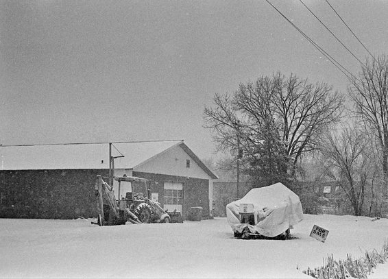 Black-and-white film photograph of a snow-covered industrial yard. A small tractor is parked near a low building in the background. In the foreground, a piece of equipment is covered with a tarp, partially buried in snow. A small “For Sale” sign sticks out of the snow to the right. Bare trees and power lines frame the scene under an overcast winter sky.