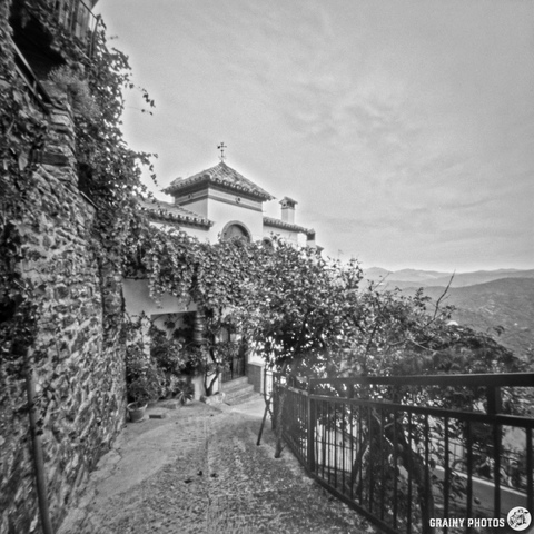 A narrow pathway leads to a charming old building covered in vines, surrounded by scenic hills in black and white. The pathway features stone walls and railings, highlighting the tranquil and rustic atmosphere of the area.