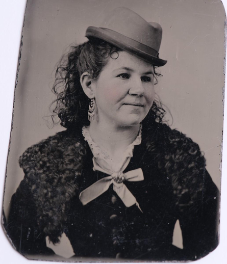 An upper body photo of a young white lady on metal. She looks to the right and has a pleasant countenance. Her dark hair is shoulder length and curly. One fairly long round earring with dangles is visible. On her head she wears a small fedora style hat that is tipped forward and kicked to the right. She wears a dark dress that's visible down to her thin waist. Over her shoulders is some sort of curly rug-like shawl. There's a light colored bow-tie at the top of the dress. 