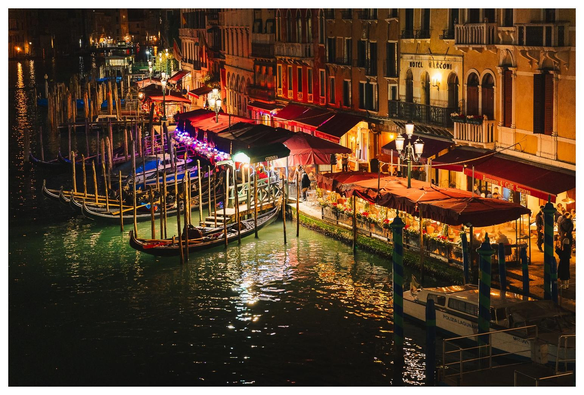 Blick von der Rialtobrücke in Venedig über den Canal Grande hin zu einigen Restaurants und Gondeln. Es ist Nacht, die Szene wird von wenigen Lampen an Häusern, Fenstern und den Außenbereichen der Restaurants erleuchtet.