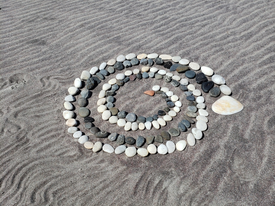 A spiral made of graduated size light and dark stones, starting at a orange-red stone and spiralling to a clamshell. It's on top of dark red sand (garnet) with wind and finger-raked ripples.