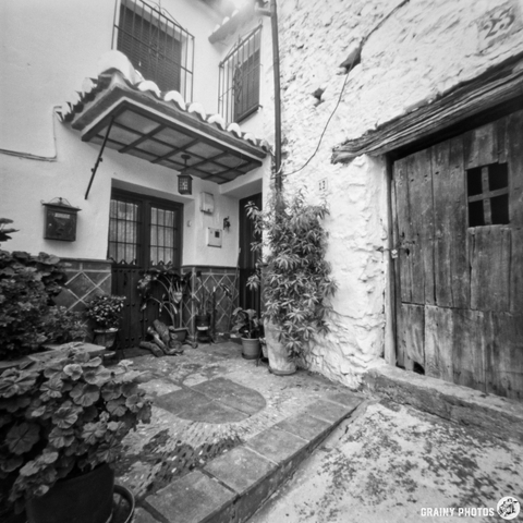 A charming courtyard entrance featuring potted plants, a stone pathway, and a rustic wooden door. The walls are textured and white, with hints of greenery, creating a welcoming atmosphere in a quaint setting.