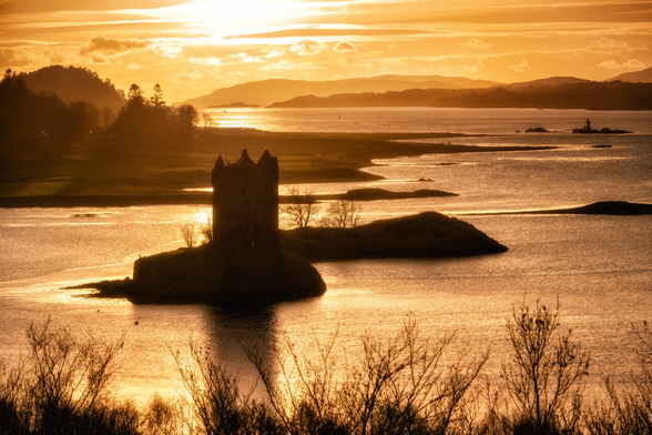 Die historische Burg Stalker präsentiert sich als markante Silhouette vor dem goldenen Licht eines Sonnenuntergangs über dem Loch Laich in Schottland. Die Wasseroberfläche reflektiert intensiv die warmen Farben des Himmels, während im Hintergrund sanfte Hügelketten in Dunst gehüllt sind. Die Komposition betont die Kontraste zwischen den dunklen Vordergrundelementen und dem leuchtenden Horizont.