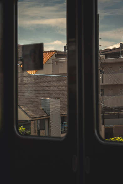 A mildy sepia coloured image taken inside a train in Osaka. The photo shows several houses against a blue sky through the blurry train doors. 