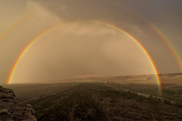A color landscape photo of a rainbow over a desert canyon area. A river is on the right. In the lower left is a small part of a cliff edge. A flat flood plain dotted with parallel lines of shrubs extends out from the foreground to a low canyon wall in the distance. A high arched rainbow extends from the river on the right to the flood plain on the far left. Parts of a second rainbow are seen above the main rainbow on the left and right. The light is much brighter under the main rainbow than above it.