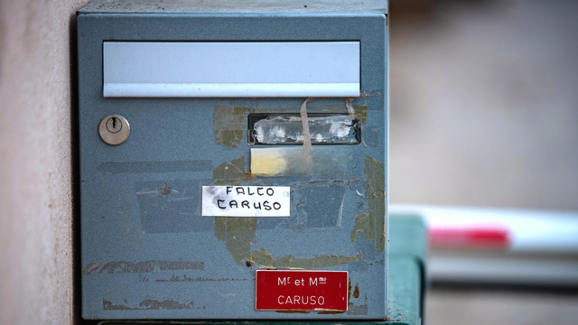 Das Bild zeigt einen grauen, an einer Wand befestigten Briefkasten in Nahaufnahme. Der Briefkasten weist deutliche Gebrauchsspuren und abgeblätterte Farbe auf. Im oberen Bereich befindet sich ein horizontaler Schlitz mit einer silbernen Abdeckung. Links davon ist ein Schlüsselloch erkennbar. Auf der Vorderseite sind zwei separate, beschriftete Etiketten angebracht. Ein weißes Etikett in der Mitte nennt "FALCO CARUSO", während ein kleineres, rotes Etikett im unteren Bereich "M. et Mme CARUSO" ausweist. Der Hintergrund ist unscharf, im Vordergrund unten rechts ist ein Teil einer rot- weißen Schranke zu sehen.