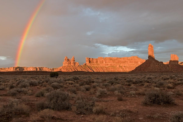 A color landscape photo of a flat sagebrush covered plain with jagged sandstone spires in the distance. The sun had just broke over a mesa top and was lighting the sandstone features across the plain. Part of a rainbow juts up on the left and disappears out of the frame. The sky is covered in dark clouds.