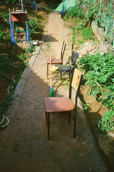 좁은 산길 가운데 빈 의자 세 개가 나란히 놓여있다. 가운데 파이프 의자에는 빗자루와 삽이 걸쳐있다. 산길 양옆으로는 작은 밭이 나있다.
