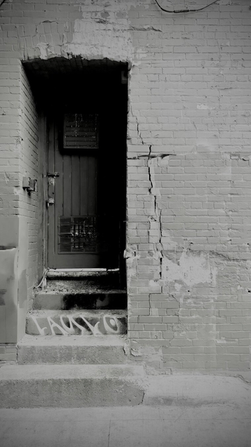 Wooden doorway recessed in a brick building, 5 stairs up to the door, 2 before the building and 3 in the door recess/alcove. Black and white. Doorway is dark but some panels are still visible on the door. Brick is deteriorating. Composition is offset; door is on the left side of the photo.