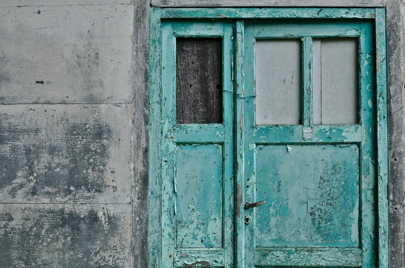 Photographie géométrique, sans profondeur d’une porte vert délavé et écaillé, fermée à l’aide de fil de fer bleu, dans un mur de béton gris.