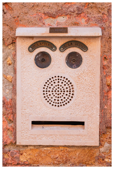 Ein in einer Mauer eingelassener Briefkasten mit Namensschild, Klingel-Drücker, gegensprechanlage und Briefschlitz. Die versch. Elemente sind so angeordnet, dass ein verblüfft/vorwurfsvoller Gesichtsausdruck entsteht.