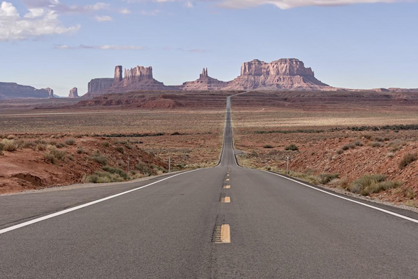 A color landscape photo looking down a long straight two lane road in a desert landscape. The landscape is reddish colored and dotted with light green sagebrush. The landscape on both sides of the road is shadowed by clouds. In the distance are several jagged blocky highly eroded sandstone formations. A couple of the large sandstone blocks have eroded into thin tall spires. Morning sunlight is breaking through and lighting the formations. The sky over the formations is mostly blue but clouds can be seen at the top and mostly in the upper left. 