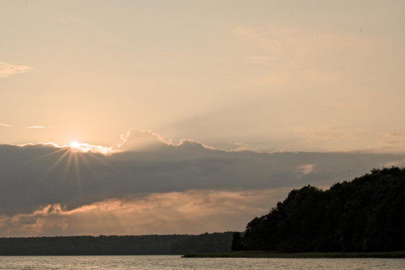 Sonnenuntergang hinter einer Wolkendecke über einem See mit bewaldetem Ufer rechts