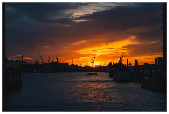 Blick zwischen zwei Bauwerken hindurch in den Hamburger Hafen. Der Himmel ist dramatisch rot/orange/blau gefärbt mit dunklen Wolken. Gebäude, Schiffe und Kräne im Hafen sind als Scherenschnitte erkennbar. Auf dem Wasser glitzert die untergehende Sonne.