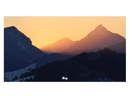 A mountainous winter landscape during sunset, the sky is completely filled with an intense, bright orange/pink glow. Silhouettes of mountains, both in the shade (in the foreground) and in/behind the glow.