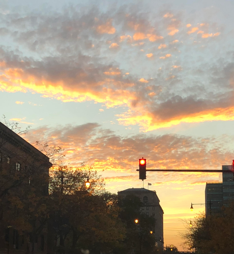 The photo shows the tops of some trees and buildings completely in shadow. Sticking out on a bar from the left in the middle of the photo is a stoplight and the red light is glowing. The rest of the photo above that is light blue sky with many clouds and the sun is starting to set and shine through the clouds, lighting up the bottom edge of the clouds into a fiery yellow and beautiful light orange. 