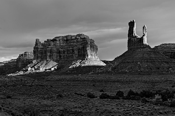 A black and white landscape photo of a highly eroded sandstone feature. The tall shear hills with conical shaped eroded debris at their base sit in a undulating sagebrush plain. The feature angles across the frame from center-right and disappearing off the center-left. On the far right is a vertical twin spire like a two prong fork. The prong furthest right is narrow and short while the prong on the left is wider and taller. Behind the feature on the right is a second blocky sandstone mass that looks like a large building or apartment complex with a lot of eroded material surrounding its base. A beam of sunlight strikes the feature on the far right and angles down lighting the base of the large block on the left. Otherwise the scene is dark and the sky is filled with dark clouds.