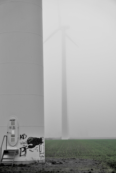 Das Foto zeigt den unteren Teil eines großen Windradturms in einer ländlichen Umgebung. Der Turm ist hellgrau und hat einige Sicherheitsaufkleber sowie ein ovales Metallgehäuse mit Belüftungsschlitzen. Auf der Oberfläche des Turms sind Graffiti-Sprayereien zu sehen. Im Hintergrund erkennt man die Rotorblätter des Windrads, die sich im Nebel oder Dunst verlieren. Vor dem Turm erstreckt sich ein grünes Feld, das vermutlich landwirtschaftlich genutzt wird. 
 ------ 
The photo shows the lower part of a large wind turbine tower in a rural setting. The tower is light grey and has several safety stickers and an oval metal housing with ventilation slots. Graffiti can be seen on the surface of the tower. In the background, the rotor blades of the wind turbine can be seen, disappearing into the fog or haze. In front of the tower is a green field, which is presumably used for agriculture.