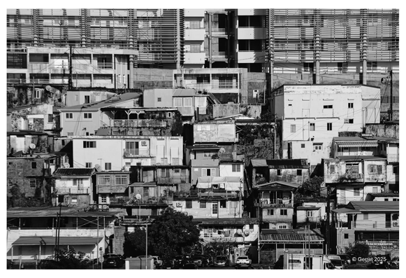 Black and white image of densely packed hillside houses in front of modern buildings. The contrast highlights urban disparity. Somber and thought-provoking.