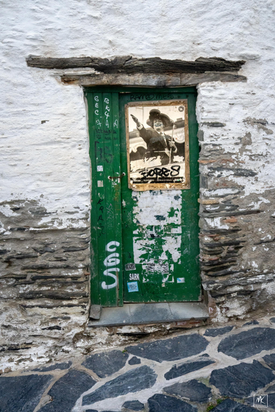 Color photo of a green door in a white painted stone wall on a stone paved street. On the door is a stained & graffitied poster with a photo of Salvador Dalí with a hand outstretched and a large odd-looking hat on his head. 