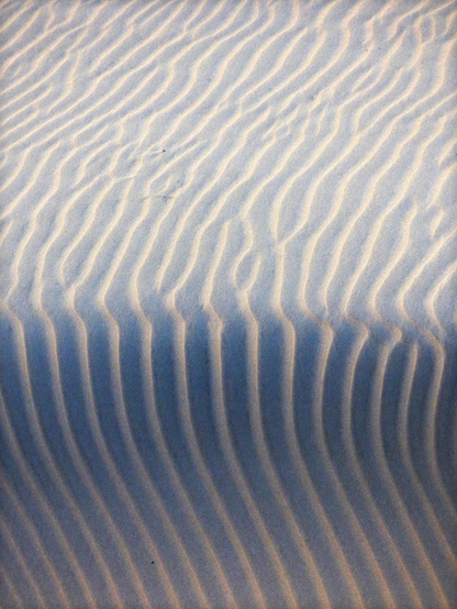 A colour photograph showing rippled sand transitioning from light into shadow. The vertical ripples pass from light into bluish shadow, splitting the frame in half.