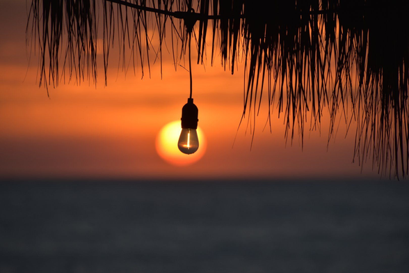 Sun setting low over the pacific ocean on Playa Las Peñitas Beach in western Nicaragua blurry in background seen through a lightbulb that is in focus on the edge of the roofline of the Simple Beach Lodge.
