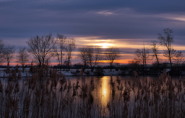 Ein Blick über einen zugefrorenen See auf das andere Ufer. Dort kann man Schnee am Ufer erkennen. Die Sonne ist schon untergegangen, beleuchtet aber noch farbenfroh den Himmel.