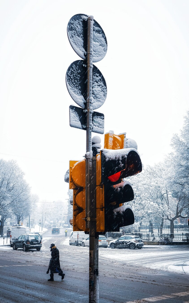 A snowy urban street scene. A traffic light ispartially covered in snow and prominently features with the red light illuminated. A person is crossing the street on the left hand side. There are several cars parked along the side of the road, also dusted with snow. In the background, trees line the street, their branches are heavy with snow, and more cars are visible further down the road.