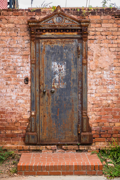 Ornate antique bank vault door set into a weathered red brick wall. The heavy metal door shows extensive rust and corrosion in shades of blue-gray, orange, and brown. The door features elaborate architectural detailing including a triangular pediment at the top with a central medallion, decorative rosettes along the frieze, and fluted pilasters on either side. Two round door handles are visible on the face of the door, which also has a deteriorating white rectangular patch. The door sits on a small brick step, and plants are growing at its base and along the top of the brick wall.