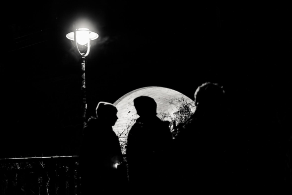 The image is a black and white photograph captured at night, showcasing a dramatic play of light and shadow. The primary focal point is a lamppost, brightly illuminated, casting a significant glow that dominates the left side of the frame. The lamppost itself has a classic design, its post appearing to be decorated with various elements.

Three silhouetted figures are positioned in the foreground, their forms defined against the bright background. They are positioned in a way suggesting casual conversation or observation.

Behind the figures, a softly lit background reveals a curved architectural structure or a bridge, adding depth to the scene. The light from this structure is diffused, and some bare branches of trees or bushes are visible in the background, further enhancing the contrast and visual interest.