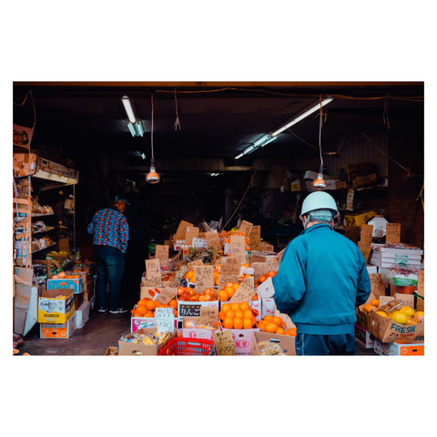 A color photo of a small fruit and vegetable shop in Nagoya. The space is crowded with cardboard boxes stacked high, each with handwritten price tags. Oranges and apples dominate the front, glowing under warm hanging bulbs. A man in a teal work uniform and helmet stands in the foreground, while another man in a colorful shirt moves about inside.