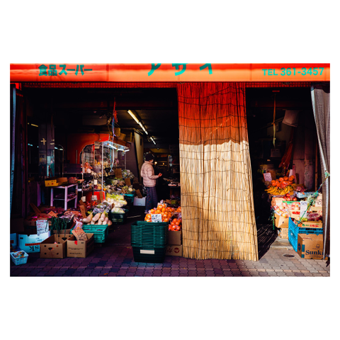 A color photo of a small produce market in Nagoya. The storefront glows under an orange awning, with a bamboo blind partially drawn to filter the morning light. Boxes of fruits and vegetables spill out onto the sidewalk—bananas, apples, oranges, leeks, and gourds. Inside, a person in a light jacket moves between the stacks.