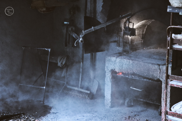 Der Blick auf einen Holzofen. Rauch vernebelt den Blick, ein bisschen Glut ist zu erahnen. Werkzeug steht nebendran. Am Bildrand ist ein Wagen mit Brotlaiben.