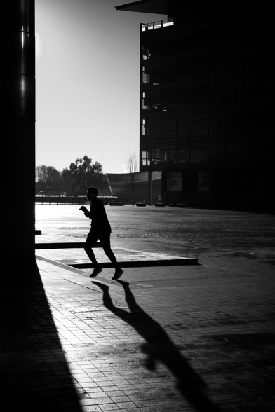 Black and white photograph of a person walking across a plaza at MediaCity in Salford Quays, creating a dramatic silhouette with a long shadow against the geometric paved surface and modern buildings.
