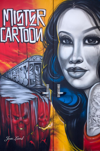 This is a colour street photo in portrait format of a double door with a bright and vivid mural of a woman and subway train in a street art style. NYC (2013).

On the right door and against a yellow/orange background is the monochrome image of a young woman, left shoulder towards the camera and looking at the lens. She has long black hair with a twirl of blue tint. Her left shoulder has an ornate, grey tattoo of a rose and some lettering that's partially in view. Her hair encroaches slightly onto the left door. The left door has a flaming red background with the words 'Mister Cartoon' in large white letters in the upper left corner. Below is a long subway, adorned with graffiti, train that disappears into the distance, it's front (or back) stops in the centre of the left door, where it meets the right door. The train is resting on multiple red, vertical wooden posts that have horrific, skull like faces near the top. 

The location: The doorway to an upmarket, downstairs bar on 5th Ave, NYC - The artwork changed regularly.
