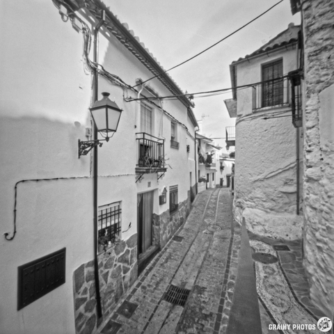 A narrow cobblestone street winds between traditional whitewashed buildings with balconies and vintage lanterns, creating a charming atmosphere in Benelauría. The scene captures a mix of urban and rustic elements in black and white.