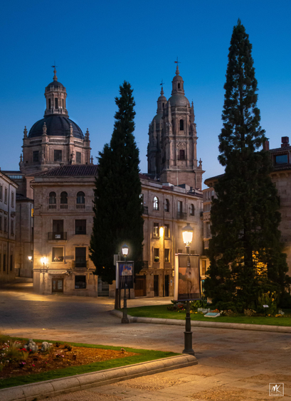 Color photo of a walkway corner in a park lit by street lamps and in the background are buildings with a baroque dome and towers lit by the pre-dawn sky. 