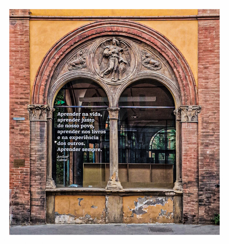 The photo shows a weathered stone building facade featuring an arched window. Above the arched window is some type of stone carved art of an angel and two lambs either side . The building is made of red bricks. The window has a quote written on it, "Aprender na vida, aprender junto do nosso povo, aprender nos livros e na experiência dos outros. Aprender sempre. Amilcar Cabral." which translates to "Learn in life, learn together with our people, learn in books and in the experience of others. Always learn. Amilcar Cabral." (I think, I don't know Spanish!)