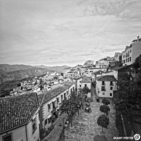A panoramic black and white view of Benelauría on a hillside, featuring whitewashed buildings, a paved plaza lined with trees, and distant mountains under a cloudy sky, capturing the serene beauty of rural architecture.